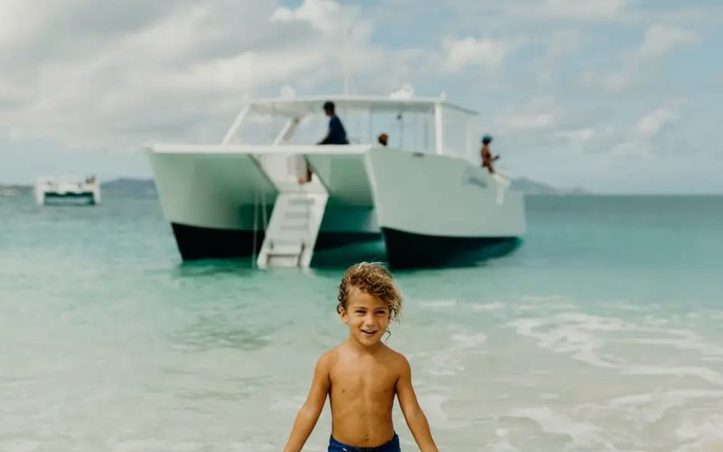 A boy on the beach at Buck Island with the Celebration catamaran anchored behind