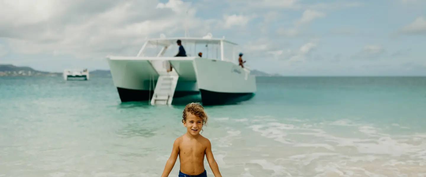 A boy on the beach at Buck Island with the Celebration catamaran anchored behind