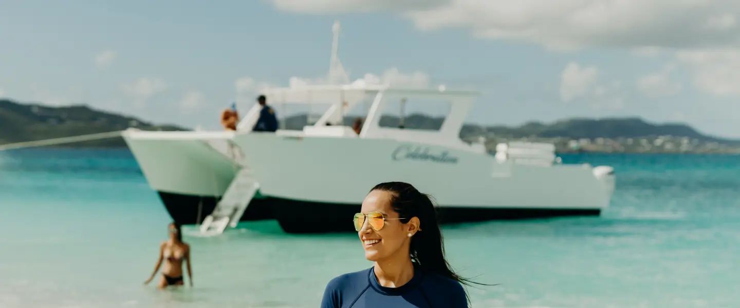 A Caribbean Sea Adventures crew member smiling in the shallows at Buck Island with the Celebration catamaran anchored behind