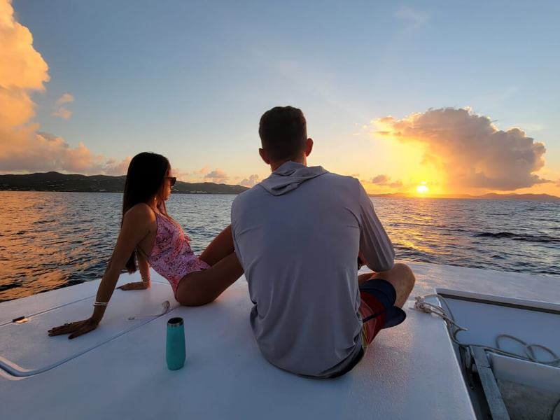 A couple sitting on the bow of Celebration as the sun sets